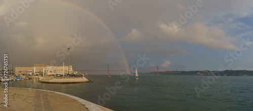 Panorama of rainbow over red bridge 25 de Abril Bridge with sailing boat during sunset, Lisbon, Portugal