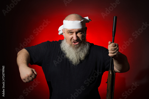 Angry old man with a black shirt with a bandage on his head with a rifle in his hand against the background of a red spot