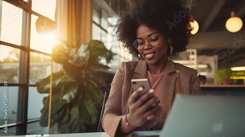 African black business woman using smartphone while working on laptop at office. Smiling mature african american businesswoman looking up while working on phone. Successful woman entre : Generative AI