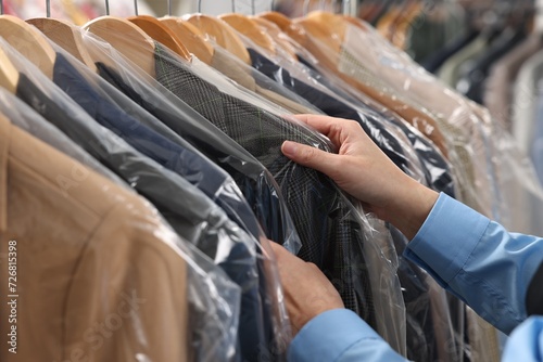 Dry-cleaning service. Woman taking jacket in plastic bag from rack indoors, closeup