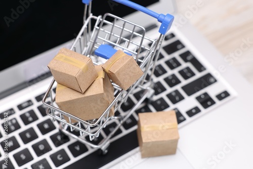 Internet store. Small cardboard boxes, shopping cart and laptop on table, closeup