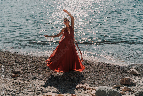Woman red dress sea. Female dancer in a long red dress posing on a beach with rocks on sunny day. Girl on the nature on blue sky background.