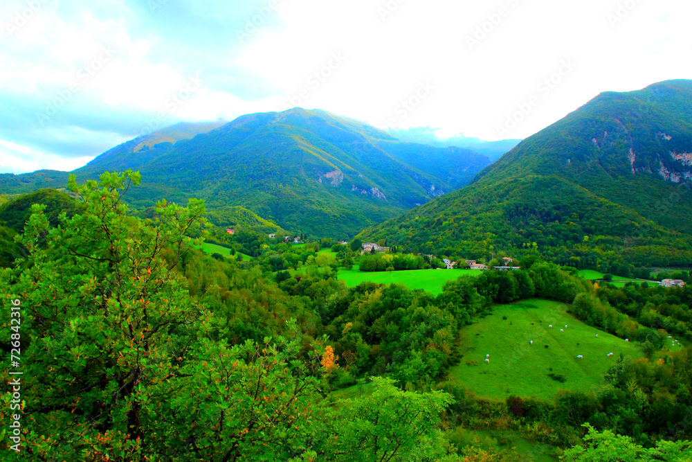 Spiritual view from Sarnano at the blue-tinted vegetation-filled Sibillini Mountains, gradiently colored from the background to the foreground, where livestock, trees and meadows combine harmonically