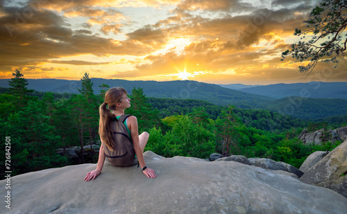 Sportive woman sitting alone taking a break on hillside trail. Female hiker enjoying view of evening nature from rocky cliff on wilderness path. Active lifestyle concept