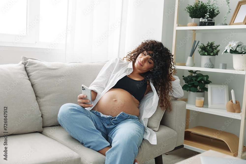Pregnant woman blogger sits on the couch at home and takes pictures of herself on the phone, selfie and video call, consultation with the doctor online, pregnancy management, freelance work