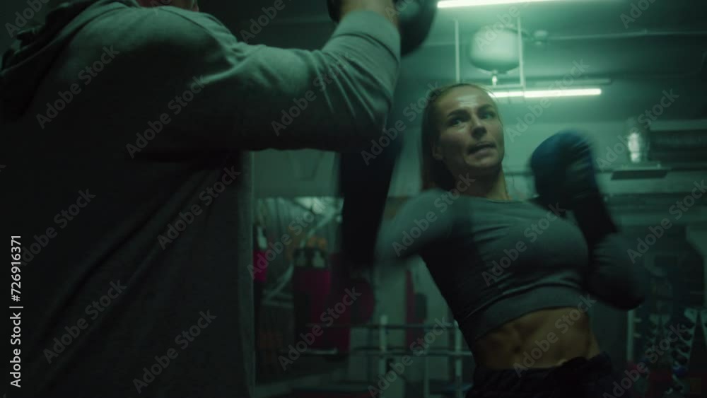 Confident female fighter in boxing gloves hits punching mitts while training in dim boxing gym. Athletic woman exercises with coach before fighting tournament. Physical activity and intensive workout. Confident female fighter in boxing gloves hits punching mitts while training in dim boxing gym. Athletic woman exercises with coach before fighting tournament. Physical activity and intensive workout.