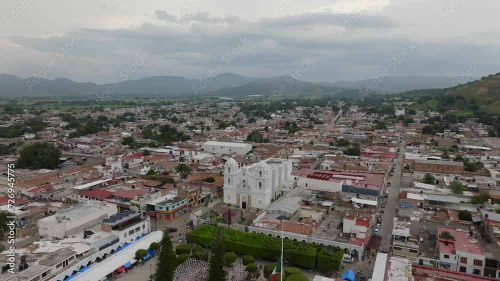 Daytime aerial view of Tecalitlan cityscape, mountains, and a green hill on the horizon. orbit right to left, wide shot
