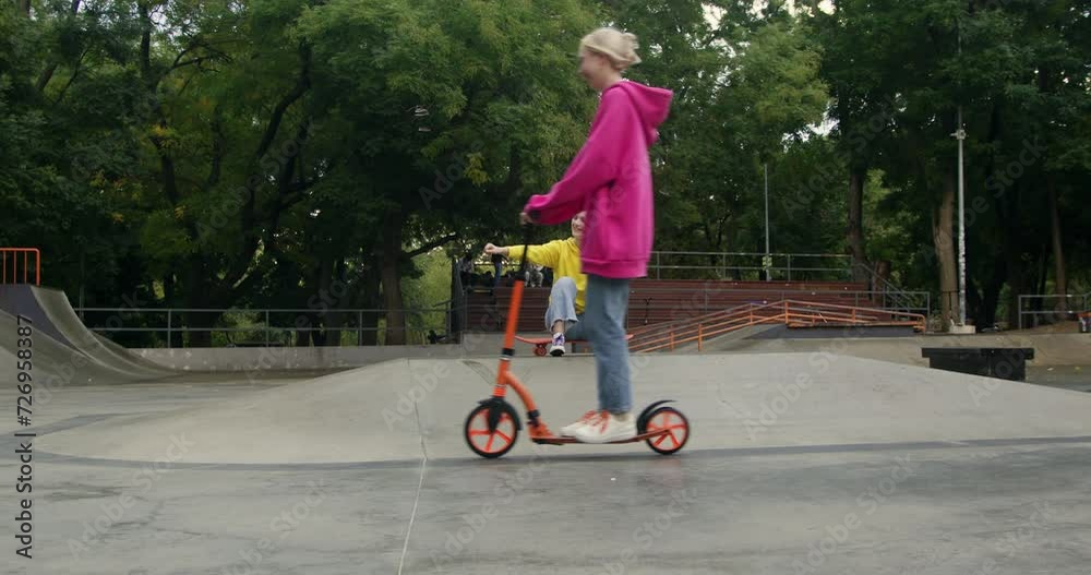 Girl sitting on skateboard cheers on blonde friend riding kick scooter. Female teenagers do extreme sports in skate park during spring weekend slow motion