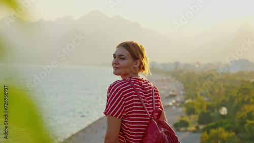 Stunnig 4K video of a white ginger girl standing  and looking to the beautiful view of the beach and mountains in the background. The video was made on a summer day in sunset (sunrise). photo