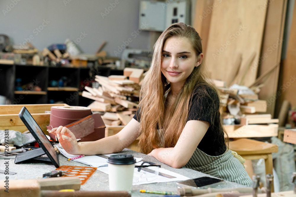 A young woman is training to be a carpenter in the workshop. She works with a laptop computer in a wood workshop. female carpenter contact customers by smartphone. SME