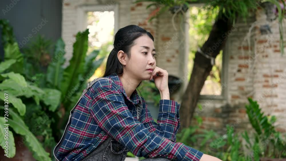 Asian female gardener Owner of small business plant shop, contemplative woman pauses in garden, surrounded by lush greenery, reflecting on botanical passions. woman in plaid takes a thoughtful moment Asian female gardener Owner of small business plant shop, contemplative woman pauses in garden, surrounded by lush greenery, reflecting on botanical passions. woman in plaid takes a thoughtful moment