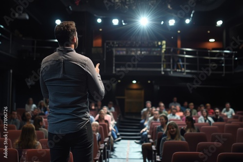 a male motivational speaker or a stand-up comedian presenting his speech in front of an audience in a microphone in a dark club or concert hall venue with red seats and selective lighting © Romana