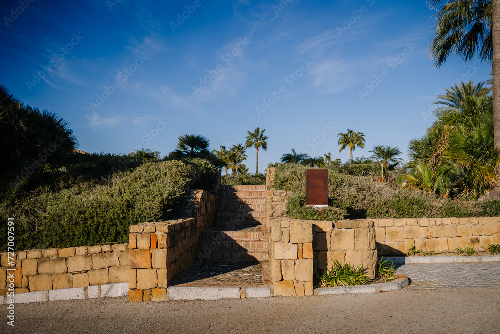 Sotogrante, Spain - January 27, 2024 - Stone stairway leading up through lush greenery, flanked by palm trees against a bright blue sky.