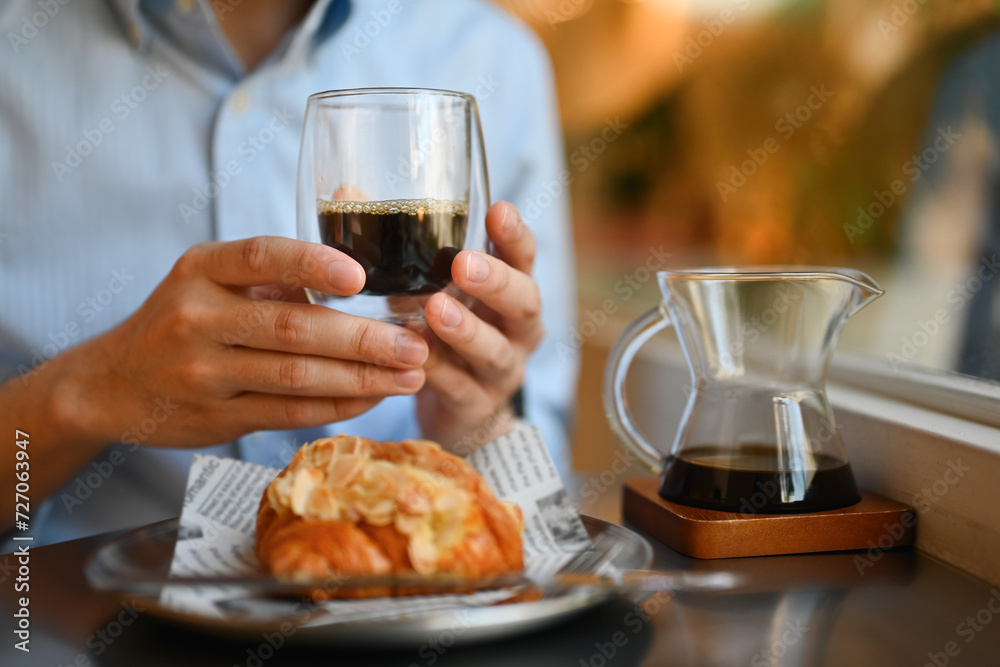 Cropped and close-up image of a Young man enjoying a black coffee and croissant beside the window