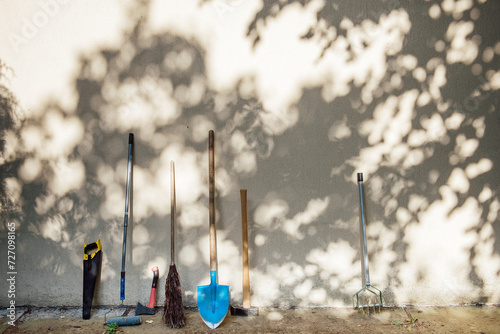 Tools for the care of the garden and the house stand near the gray wall in the shade of trees and the rays of the sun. A real life shot with space for text, banner size photo