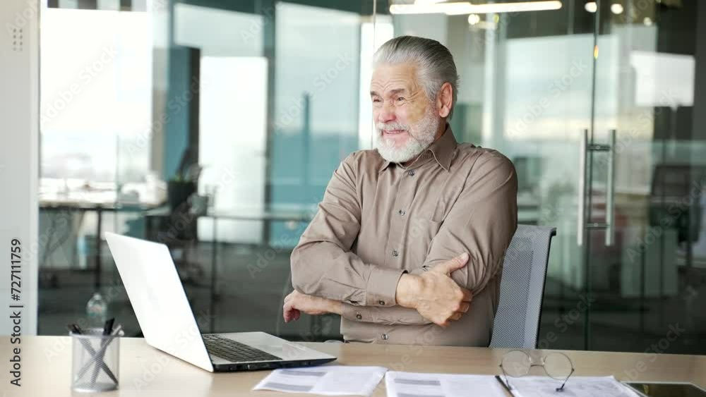 Senior gray haired bearded businessman suffering from elbow pain sitting at workplace in business office. Upset elderly male is massaging a joint, rubs his hand. He has joint injury or ligament sprain Senior gray haired bearded businessman suffering from elbow pain sitting at workplace in business office. Upset elderly male is massaging a joint, rubs his hand. He has joint injury or ligament sprain