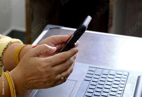 Hand of unrecognizable female in Indian outfit (Saree) using smartphone while working with laptop at home