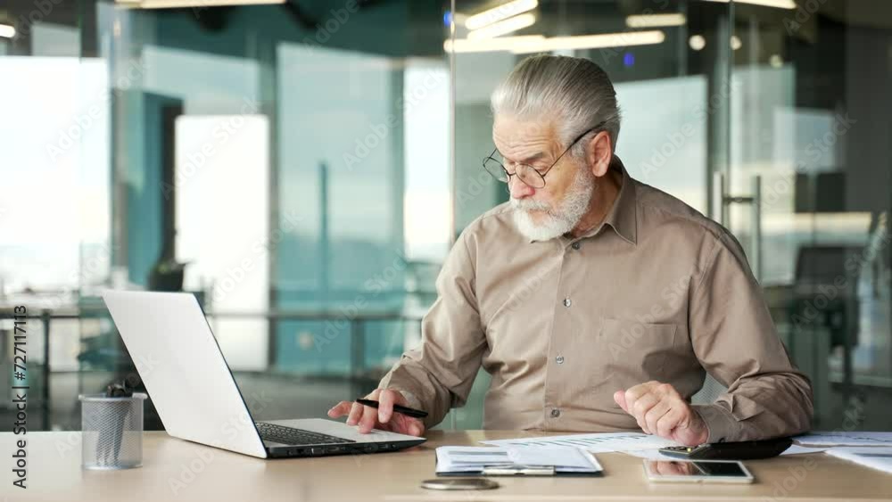 Busy senior gray haired bearded financier is doing paperwork using laptop computer sitting at workplace. Confident businessman working fills out documents writing, making tax return in business office Busy senior gray haired bearded financier is doing paperwork using laptop computer sitting at workplace. Confident businessman working fills out documents writing, making tax return in business office