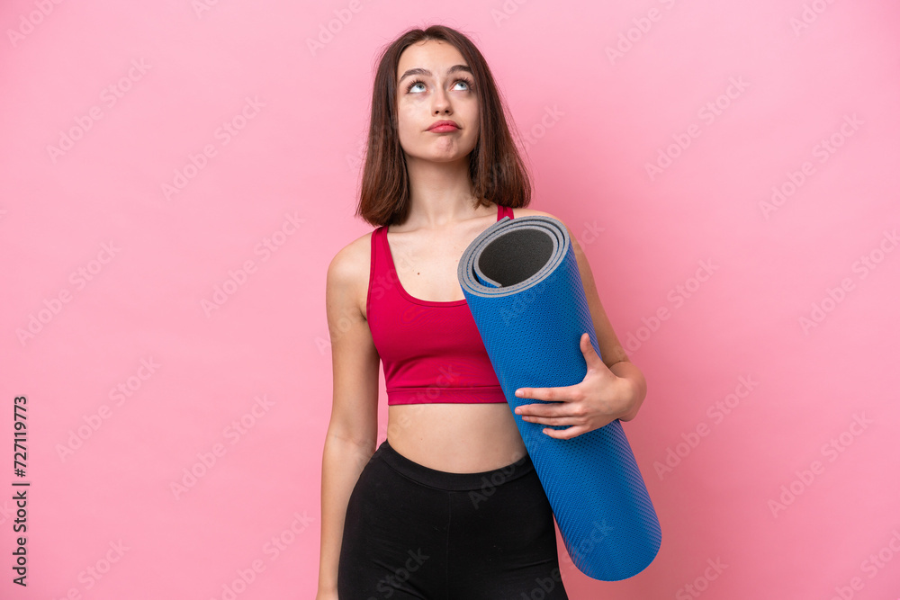 Young sport Ukrainian woman going to yoga classes while holding a mat isolated on pink background and looking up Young sport Ukrainian woman going to yoga classes while holding a mat isolated on pink background and looking up