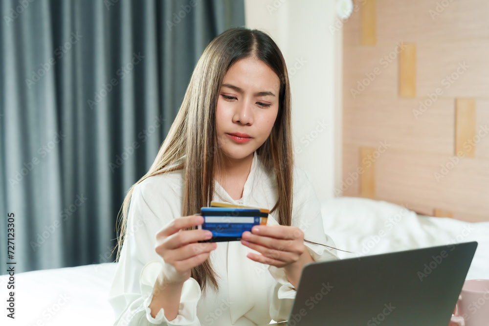 Serene young woman holding a credit card looks at her laptop, contemplating a purchase in her calm bedroom. thoughtful female with a credit card at hand navigates online shopping portal on laptop Serene young woman holding a credit card looks at her laptop, contemplating a purchase in her calm bedroom. thoughtful female with a credit card at hand navigates online shopping portal on laptop