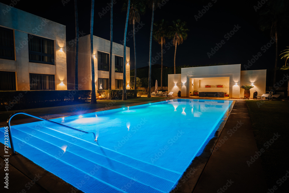 Sotogrante, Spain - January 27, 2024 - Night view of an illuminated blue pool with surrounding palm trees and a modern building with lights.