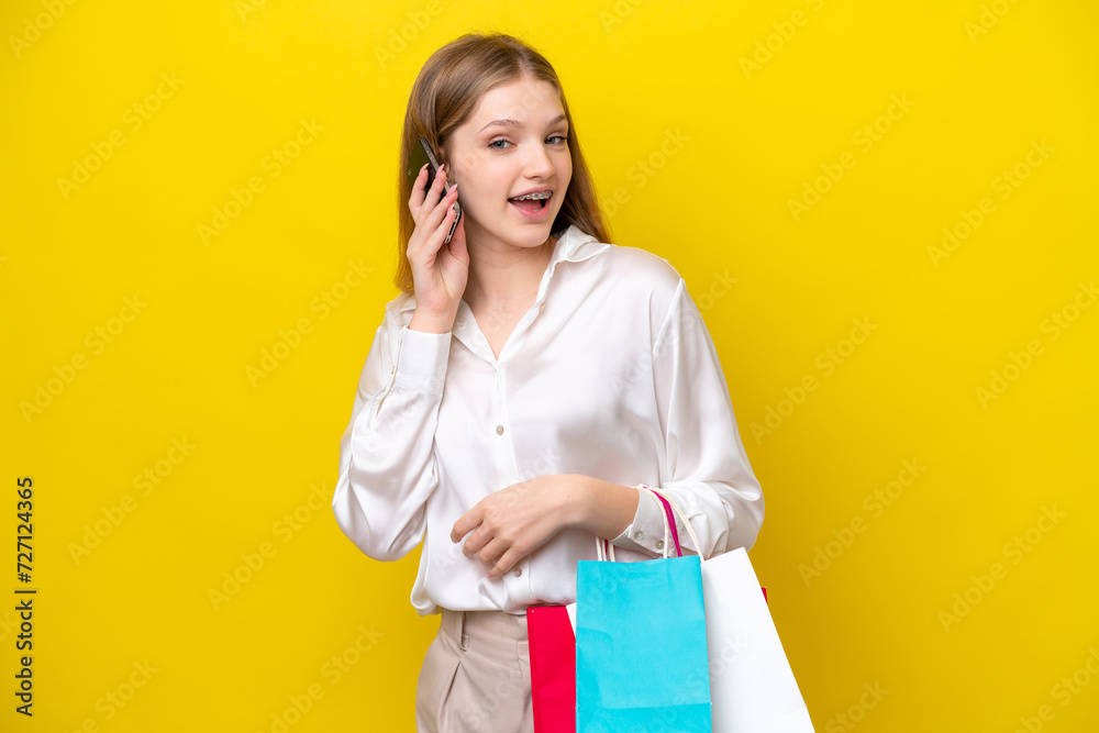 Teenager Russian girl isolated on yellow background holding shopping bags and calling a friend with her cell phone Teenager Russian girl isolated on yellow background holding shopping bags and calling a friend with her cell phone