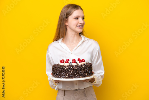 Teenager Russian girl holding birthday cake isolated on yellow background looking side
