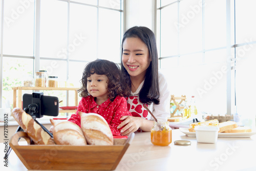 Smiling cute daughter girl sits on mother lap and uses smartphone to record social media channel blogger vlog at kitchen. Happy influencer family cooking for streaming online podcast on mobile phone.