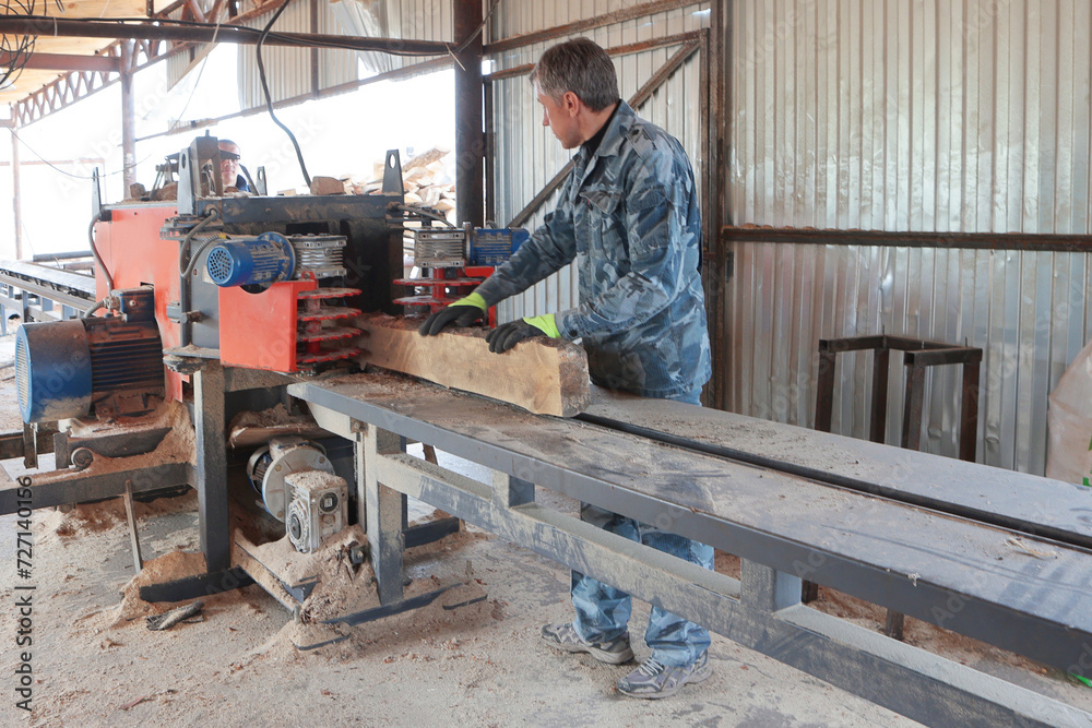 A carpenter works at a sawmill in a wood production company. The process of processing logs at a sawmill. The tree trunk is being sawed. The sawmill.