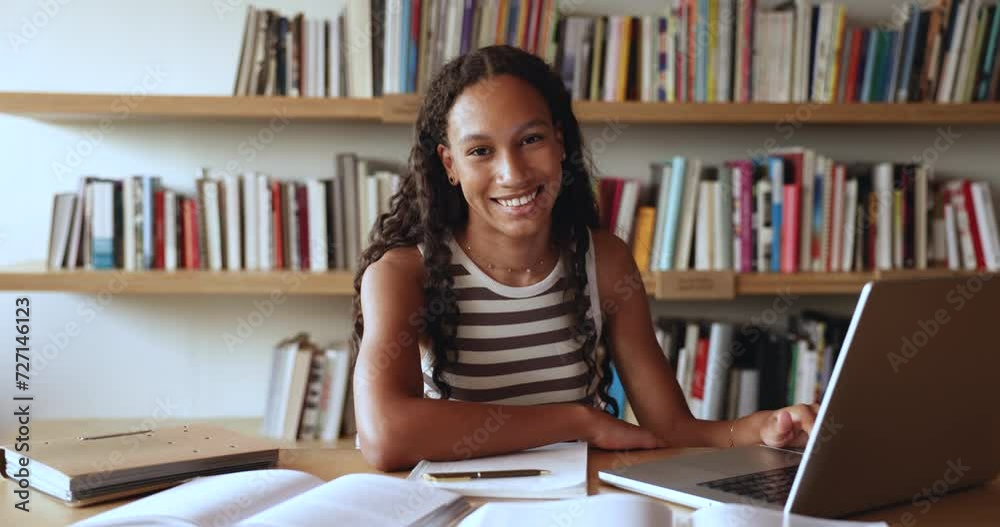 African student girl sit at desk, prepare for exam use laptop, search information, study on project in college library, do homework. Smiling girl look at camera, feel happy enjoy university education