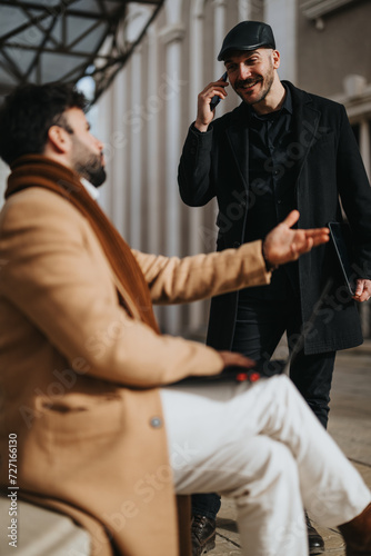 Two stylish men in winter coats having a cheerful discussion on a city street. One is on a phone call  while the other waits.