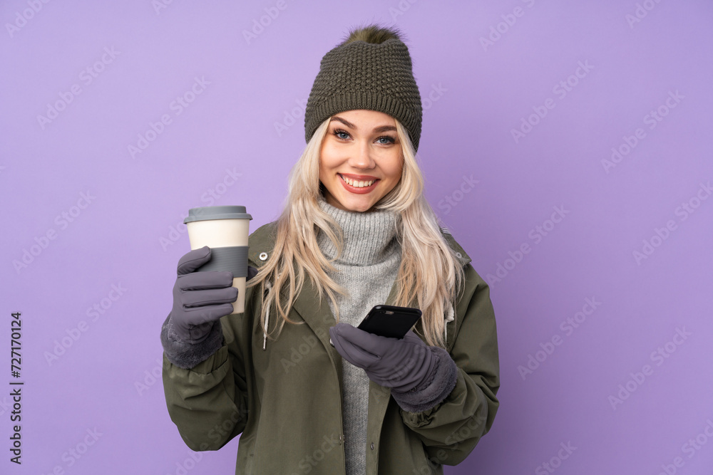Teenager blonde girl with winter hat over isolated purple background holding coffee to take away and a mobile Teenager blonde girl with winter hat over isolated purple background holding coffee to take away and a mobile