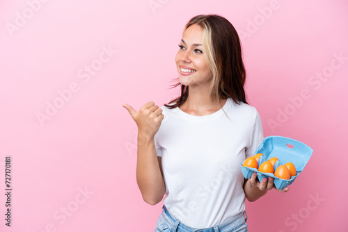 Young Russian woman holding eggs isolated on pink background pointing to the side to present a product