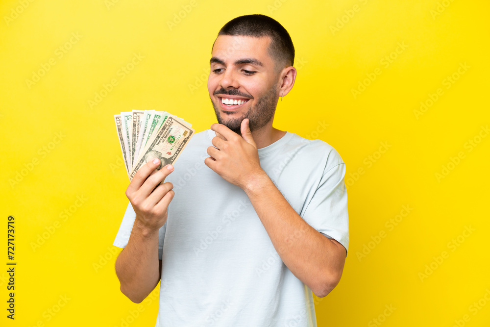 Young caucasian man taking a lot of money isolated on yellow background looking up while smiling Young caucasian man taking a lot of money isolated on yellow background looking up while smiling