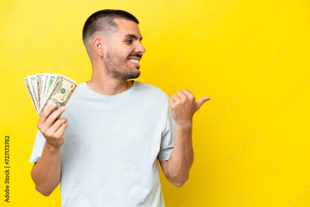 Young caucasian man taking a lot of money isolated on yellow background pointing to the side to present a product Young caucasian man taking a lot of money isolated on yellow background pointing to the side to present a product