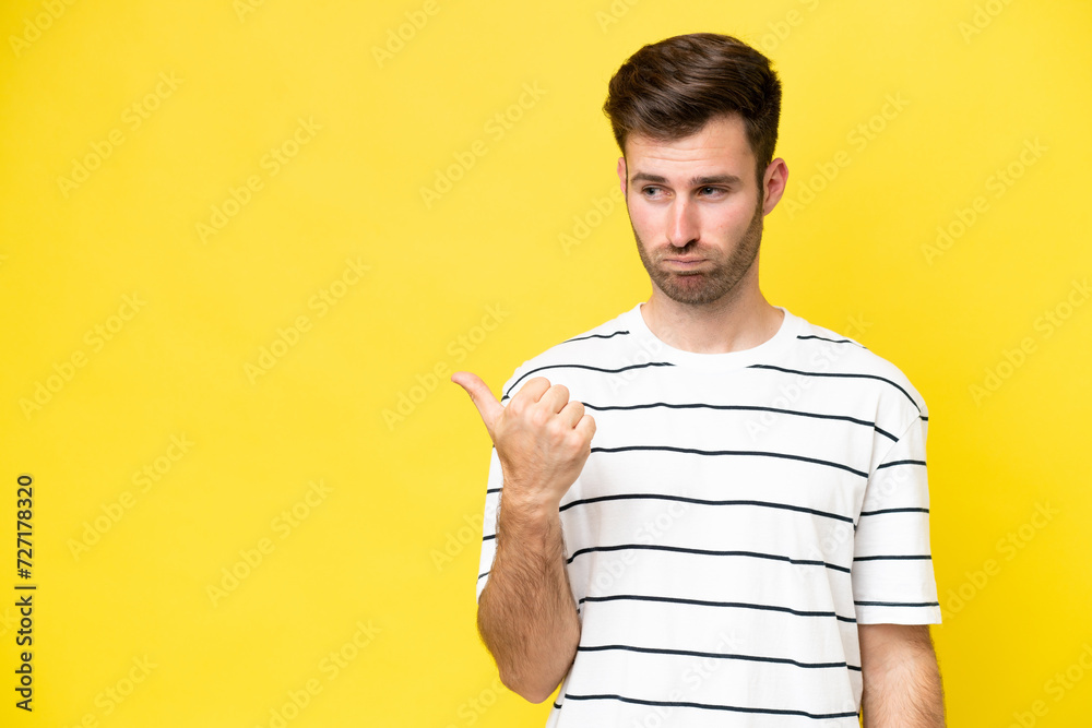 Young caucasian man isolated on yellow background unhappy and pointing to the side