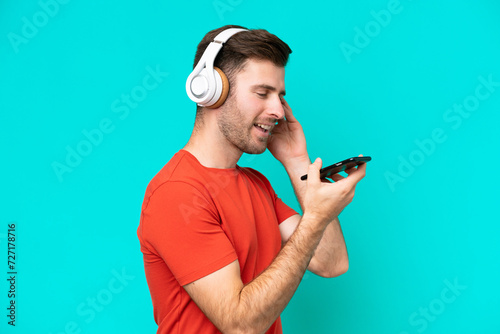 Young caucasian man isolated on blue background listening music with a mobile and singing