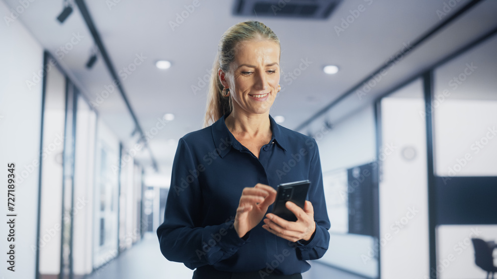 Portrait of a Smiling Adult Woman Browsing Internet and Social Networks on Her Smartphone while Walking in a Corporate Office. Young Female Marketing Manager Checking Trends and News Updates. Portrait of a Smiling Adult Woman Browsing Internet and Social Networks on Her Smartphone while Walking in a Corporate Office. Young Female Marketing Manager Checking Trends and News Updates.