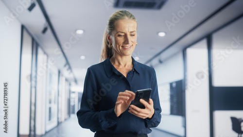 Portrait of a Smiling Adult Woman Browsing Internet and Social Networks on Her Smartphone while Walking in a Corporate Office. Young Female Marketing Manager Checking Trends and News Updates.