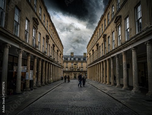 Dramatic urban street scene with historic architecture, tall columns, and moody overcast skies, as a few pedestrians walk along the cobblestone pathway.