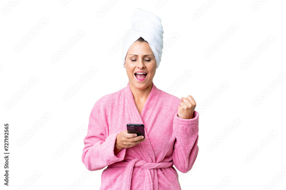 Middle age caucasian woman in a bathrobe over isolated background with phone in victory position Middle age caucasian woman in a bathrobe over isolated background with phone in victory position