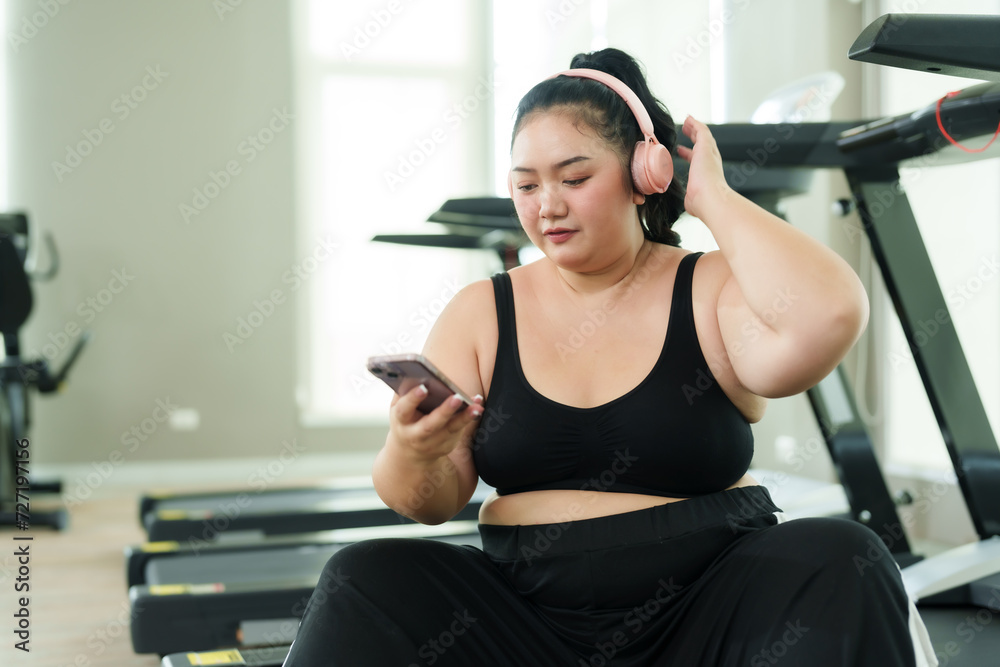 Chubby Asian woman exercise in gym, woman in activewear joyfully uses her smartphone during break, reflecting balance of leisure and fitness. Sitting comfortably on a treadmill, happy engages Chubby Asian woman exercise in gym, woman in activewear joyfully uses her smartphone during break, reflecting balance of leisure and fitness. Sitting comfortably on a treadmill, happy engages
