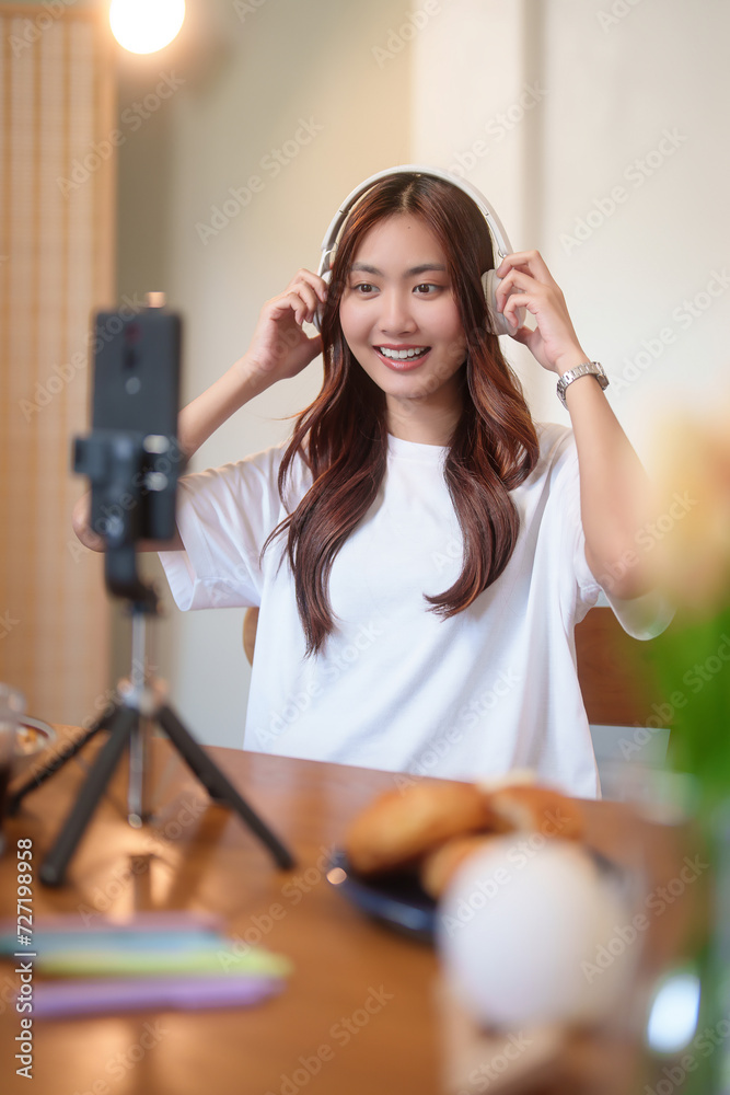Young asian vj women wearing headphone and using digital smartphone to recording video and speaking morning talks about news in live streaming on channel while working with happiness at home studio