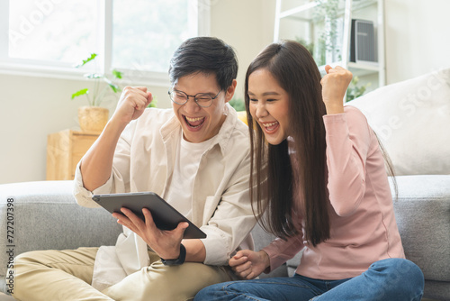 Happy excited, smiling asian young couple love using tablet pc, great deal or business success, received or getting cash back, tax refund, good news by mail while sitting on floor at home. photo