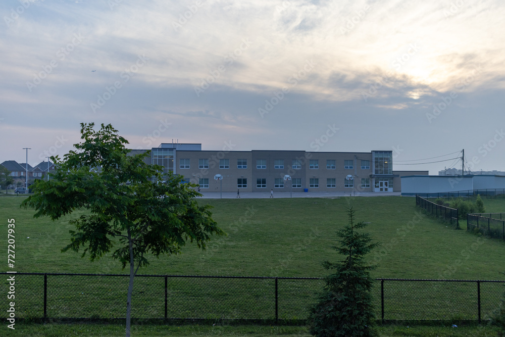 Obraz premium Modern school building - against a backdrop of a cloudy sky at dusk - surrounded by a well-maintained green lawn - bordered by a black fence. Taken in Toronto, Canada.