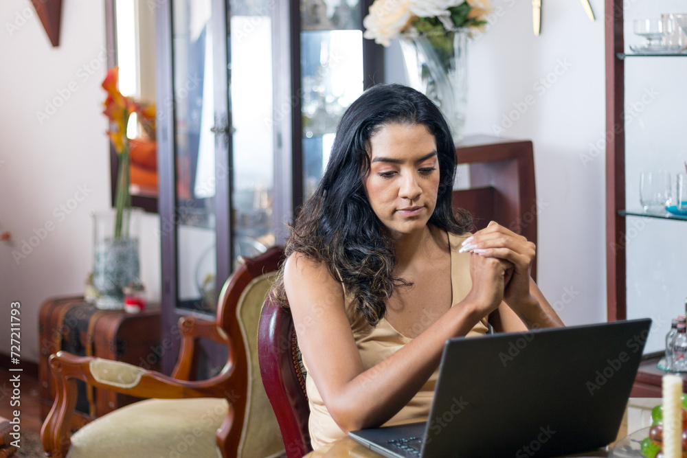 Naklejka premium Concerned black haired latin woman working on her laptop in her living room