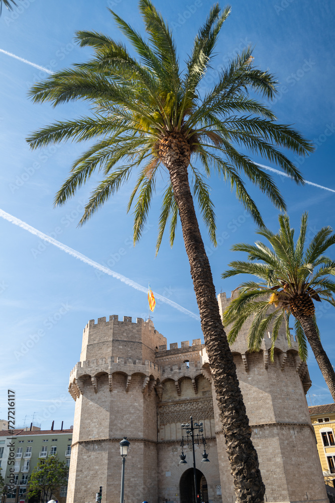 Valencia, Spain - May 12 2023 :  Torre Serranos Towers, a city wall and fortress in bright blue summer sunshine with palm tree in the foreground. Vertical mobile format layout