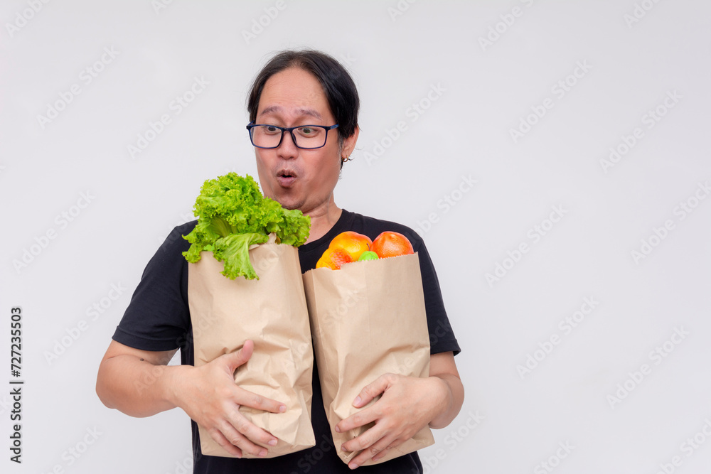 Surprised Asian man holding paper grocery bags stuffed with fresh vegetables and fruit against a white background. Surprised Asian man holding paper grocery bags stuffed with fresh vegetables and fruit against a white background.