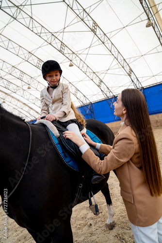 Young woman is training horse in riding hall. The girl is sitting on saddle and the woman is holding the horse's reins.