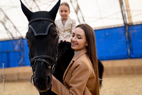 Young woman and her little daughter are walking around the arena with black horse. The concept of horse riding.
